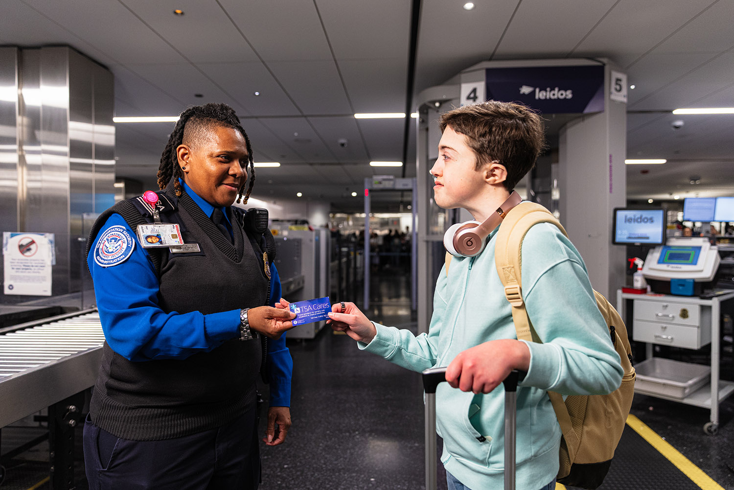 A TSA officer checks a teenager’s boarding pass at an airport security checkpoint. The teenager holds a suitcase and headphones rest around their neck.