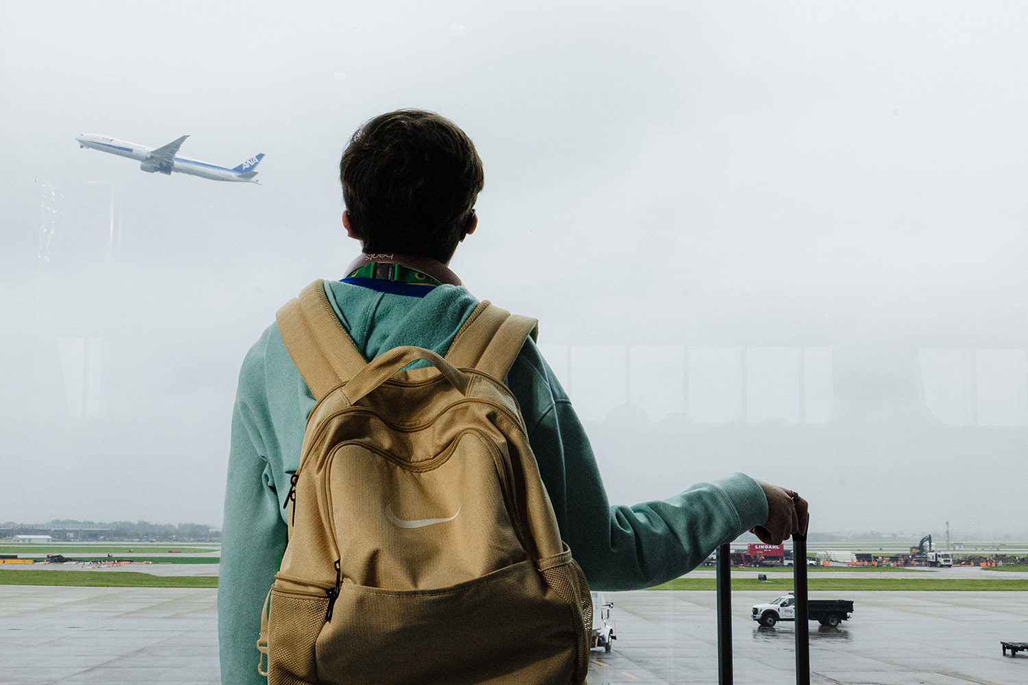 A young traveler, wearing a hoodie and a backpack, gazes out a terminal window at a departing airplane in overcast weather.