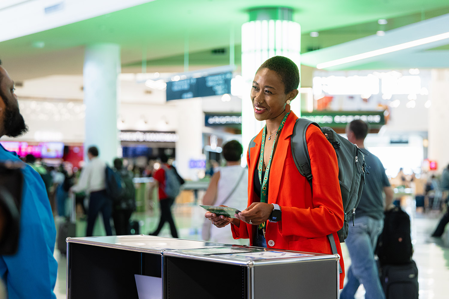 A person in a vibrant red jacket stands at an information desk in a bustling airport, holding a postcard with a busy terminal in the background.