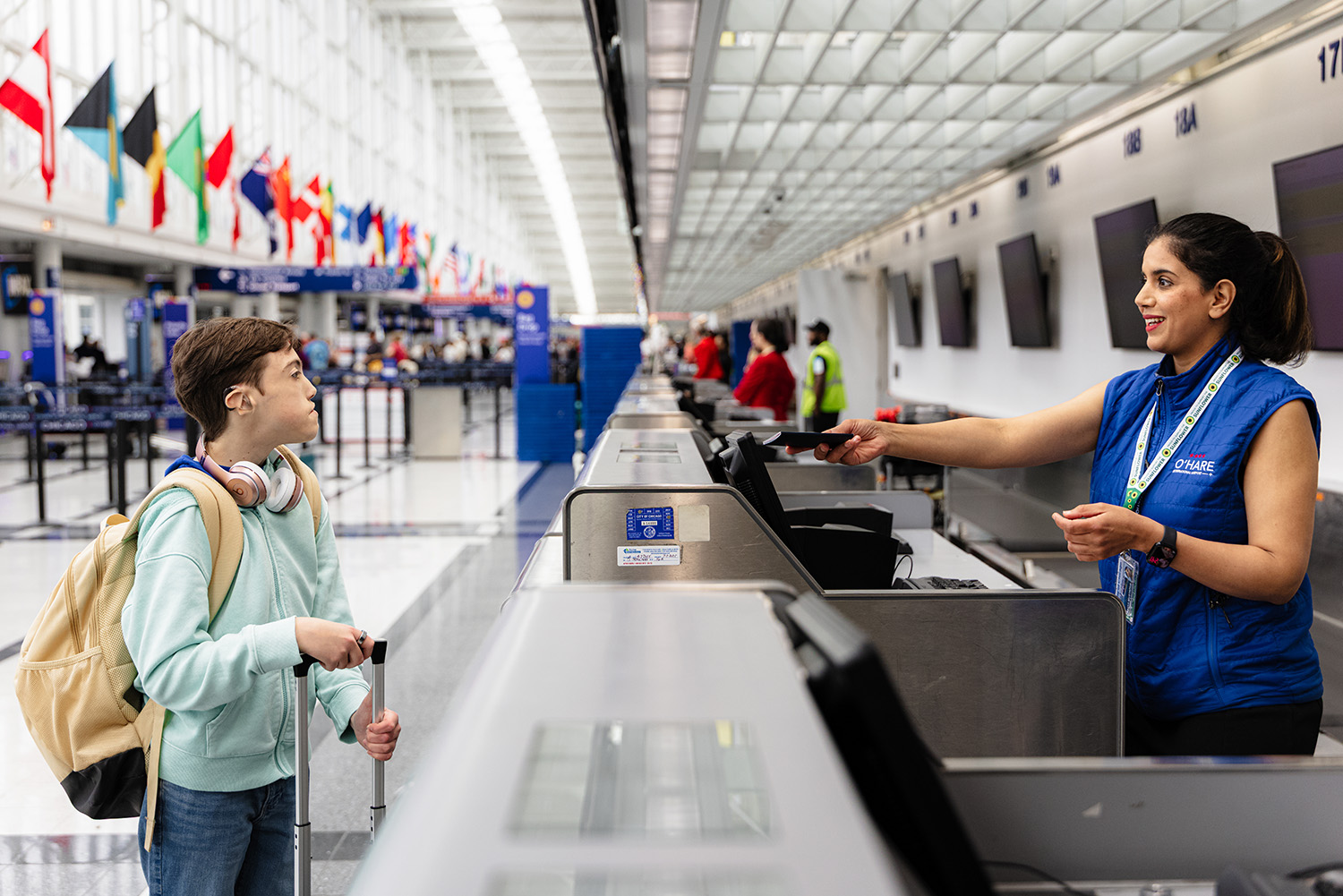 A traveler with a backpack and headphones stands at an airport check-in counter, engaging with staff amid a bustling terminal.