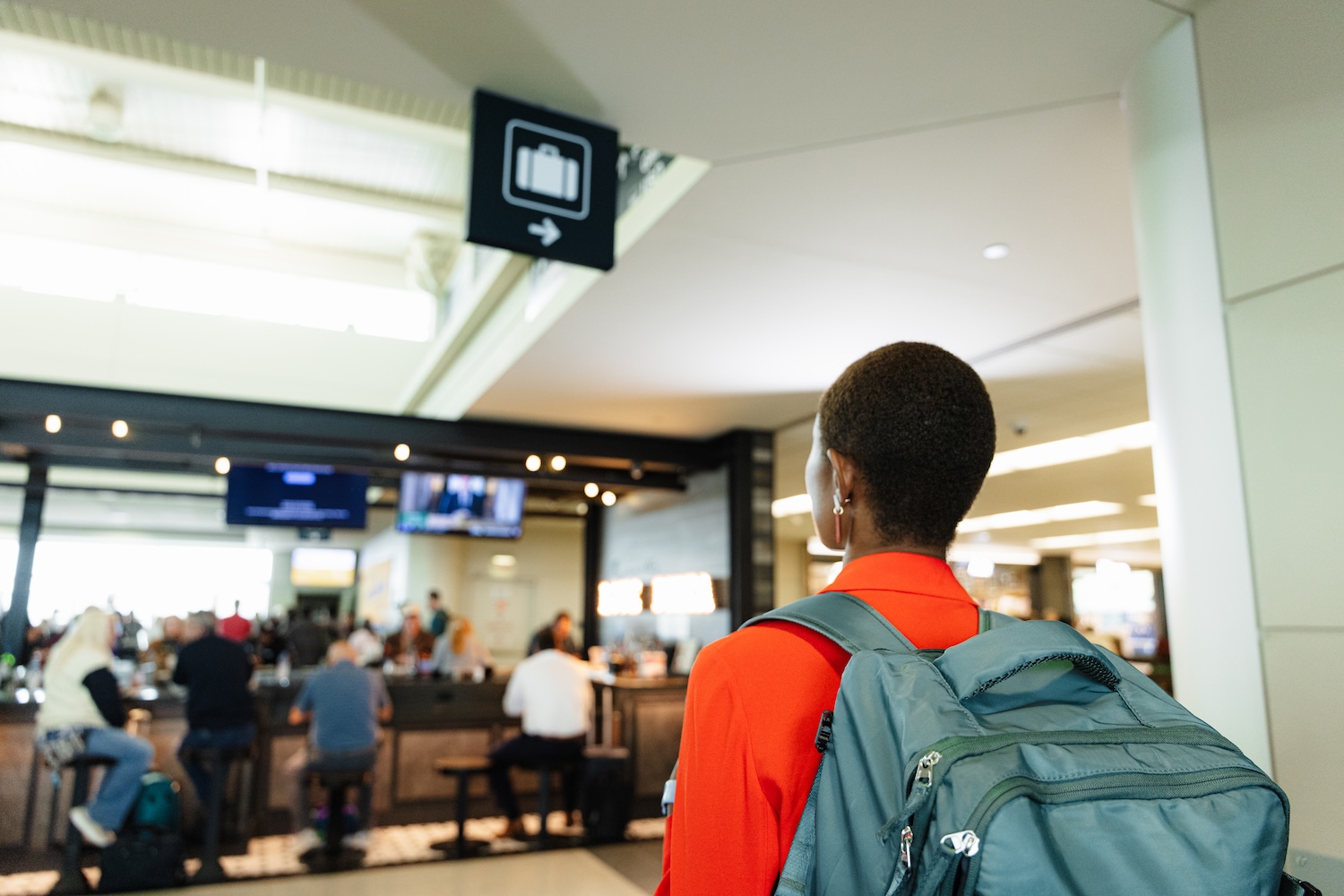 A traveler with short hair, wearing a red jacket and a teal backpack, stands inside an airport terminal looking toward overhead signs. The terminal is busy with people sitting and walking in the background. An overhead baggage claim sign is visible.