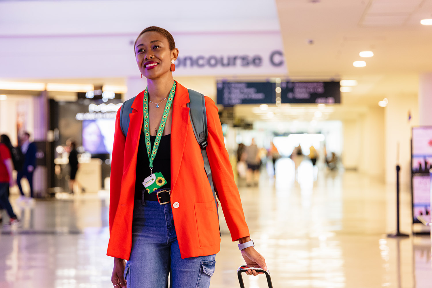 A woman wearing a red blazer and jeans walks through an airport terminal, pulling a suitcase and smiling, with people and signs in the background.